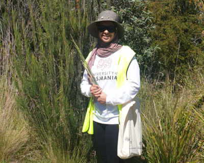 Image of Ashwag in a hi-vis vest holding a plant cutting outside. She is also wearing a bucket hat and sunglasses and is smiling.