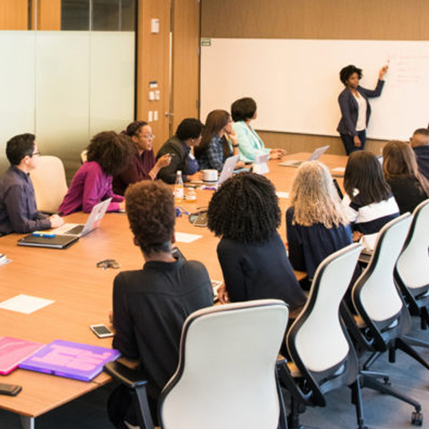 Image of a group of people in a meeting room sitting and listening to a women at a white board explaining something