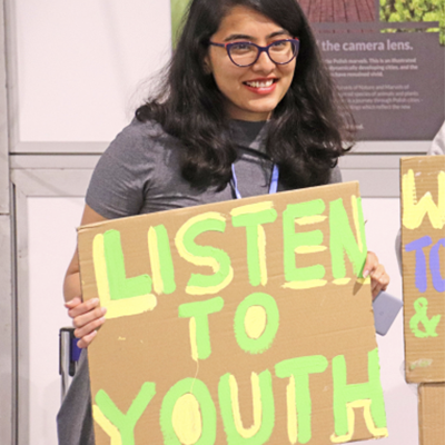 Image of Sanja smiling for the camera holding up a cardboard sign painted saying "Listen to Youth".