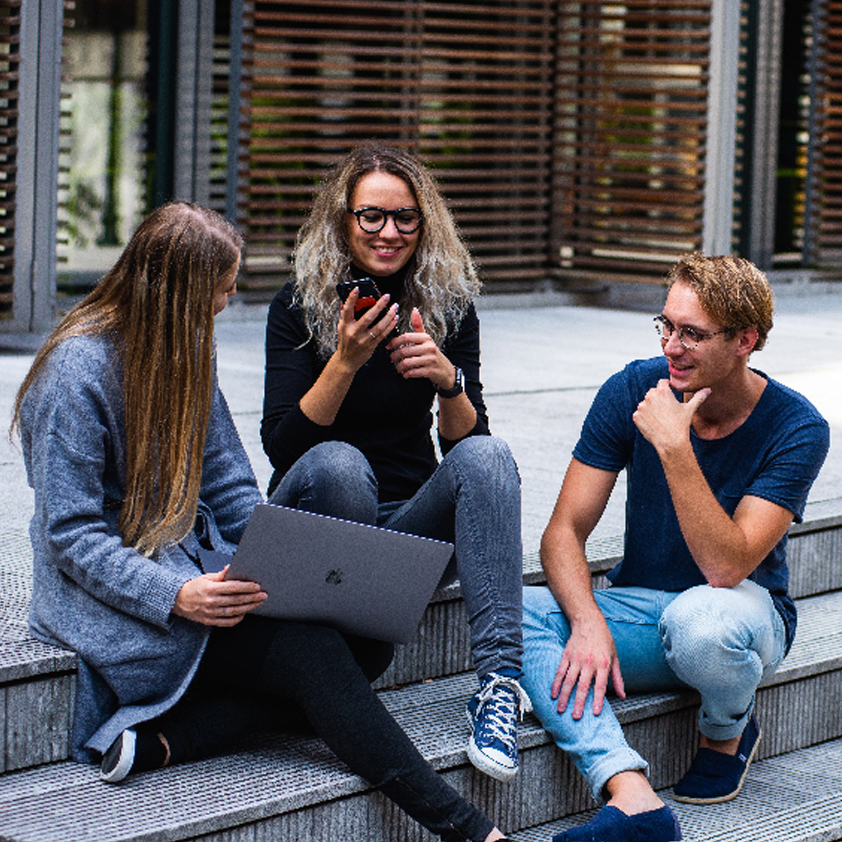 Image of three young adults sitting on the steps of a university in casual conversation.