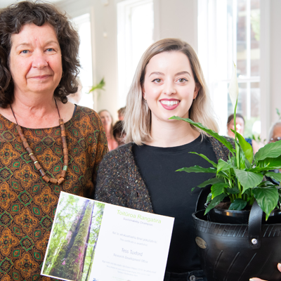 Image of Tess Tuxford receiving a certificate. Tess is smiling and holding the certificate in one hand and a green plant in a black pot in the other.